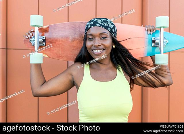 Smiling woman wearing headscarf holding skateboard in front of wall