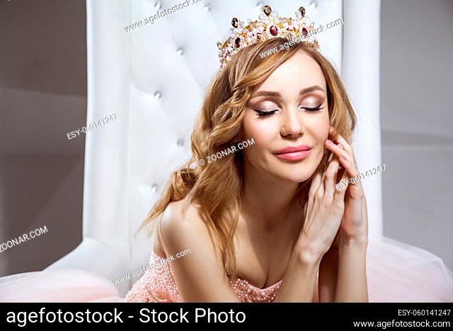 Portrait of beautiful young woman in crown and luxurious pink dress posing on stylish white armchair. sitting, touching her face and smiling with closed eyes