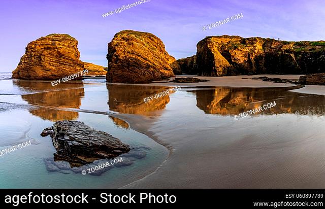 A peaceful sunset on a sandy beach with tidal pools and jagged broken cliffs behind