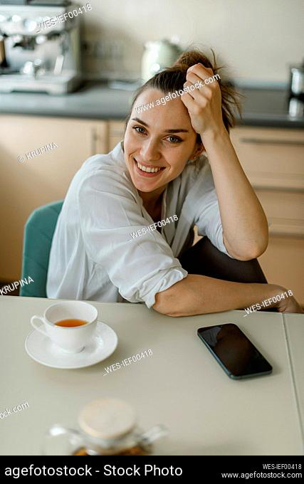 Smiling woman sitting on table with smart phone and tea