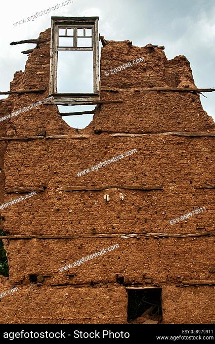 Brick wall with adobe clay plaster and broken wooden window frame remained of ruined old rural country house on cloudy blue sky background