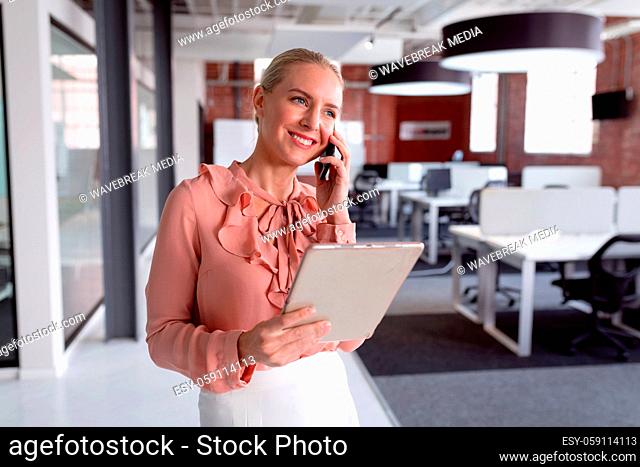 Happy caucasian businesswoman standing in office using tablet, talking on smartphone and smiling