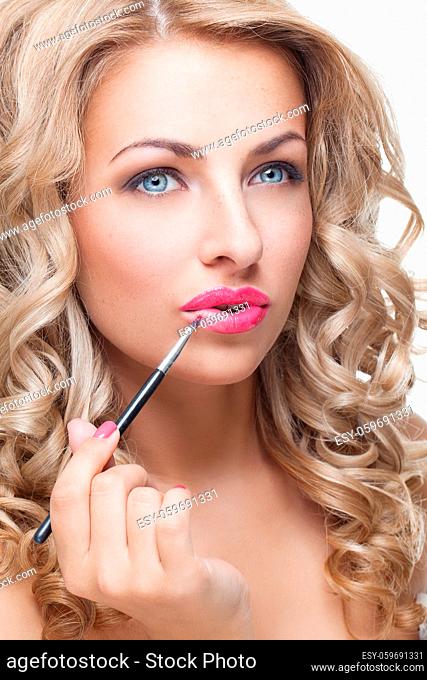 Beautiful young woman with long blond curly hair putting pink lipstick on with lip brush. Isolated on white background. Copy space
