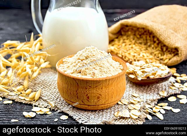 Flour oat in a bowl, milk in a jug, oatmeal in a spoon on burlap, grain in bag, oaten stalks on wooden board background