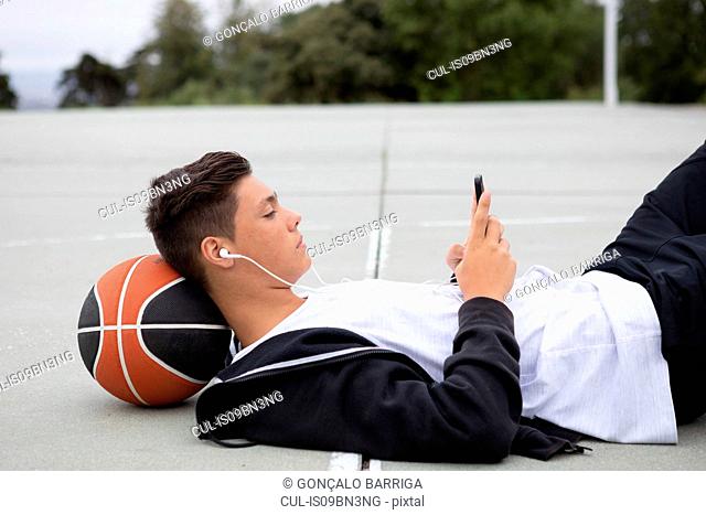 Male teenage basketball player lying on basketball court looking at smartphone