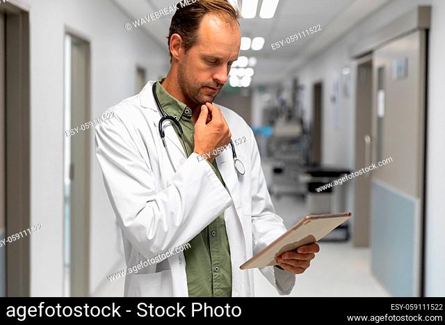 Portrait of caucasian male doctor standing in hospital corridor looking at tablet