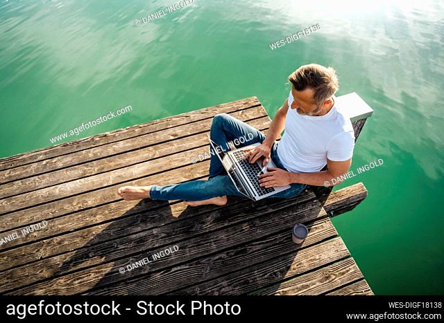 Freelancer working on laptop sitting at pier over lake