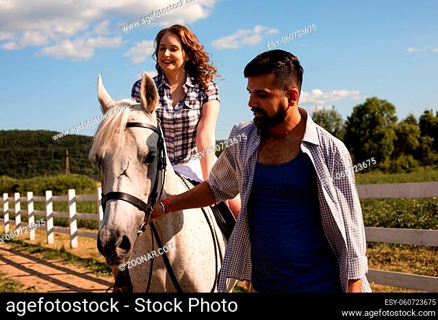 The young bearded man leads a white horse. The beautiful young woman is sitting on a horse on sunny day