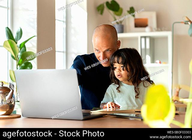Father working on laptop while sitting with toddler at home office