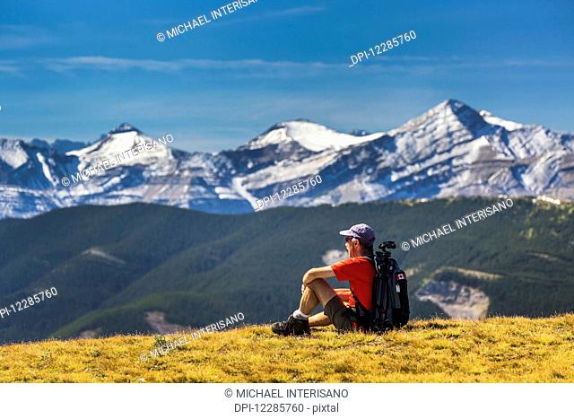 Male hiker sitting on a grassy mountain top overlooking foothills, snow capped mountain range and blue sky with clouds; Kananaskis Country, Alberta, Canada