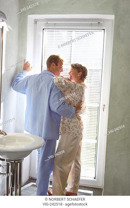 indoor, full-figure, young couple wearing pyjama stands bare feeted arm-in-arm at a glassdoor in the bathroom looking at each other  - GERMANY, 27/02/2005