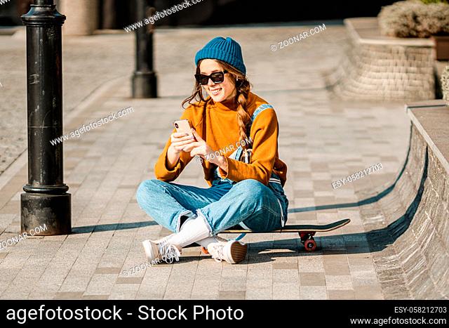 Girl sitting on skateboard and use mobile phone. Outdoors, urban lifestyle. cute skater girl sitting on skate board checking smart phone listening to music...