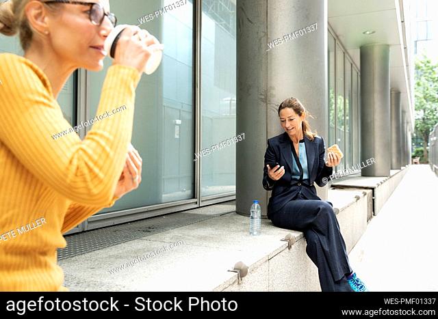 Woman drinking coffee while sitting with businesswoman using mobile phone in background in city