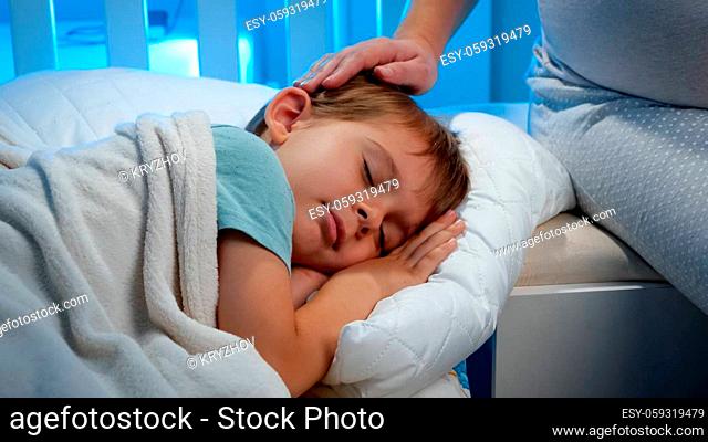 Closeup of little sleeping boy in bed and loving mother stroking him on head. Cute toddler boy sleeping while mother caress him