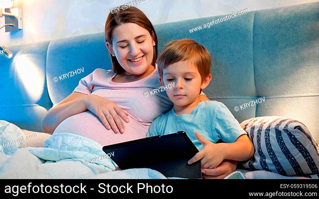 Happy smiling boy with mother watching cartoons on tablet computer in bed at night
