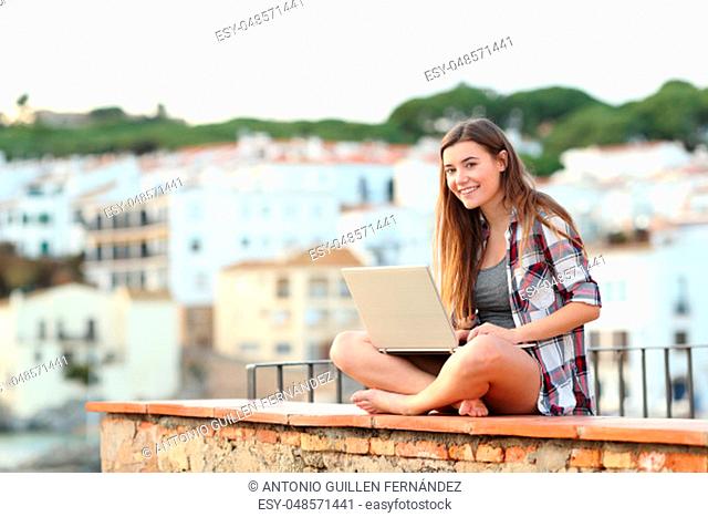 Happy teen looking at camera using a laptop sitting on a ledge in a coast town on vacation