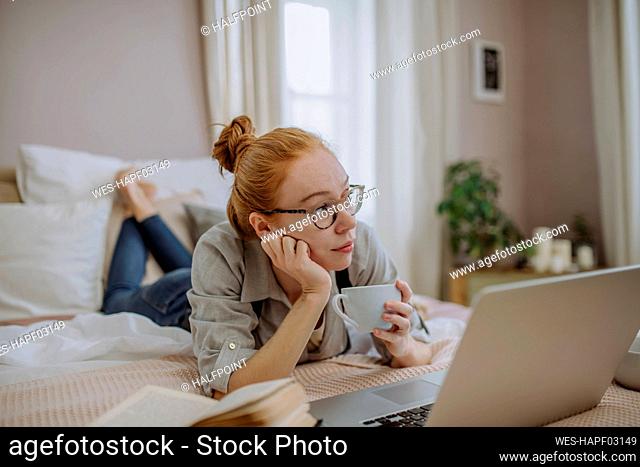 Thoughtful woman holding coffee cup lying with laptop on bed at home