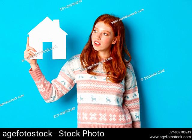 Real estate concept. Image of cute redhead girl looking curious at paper house model, thinking of buying property, standing in sweater against blue background