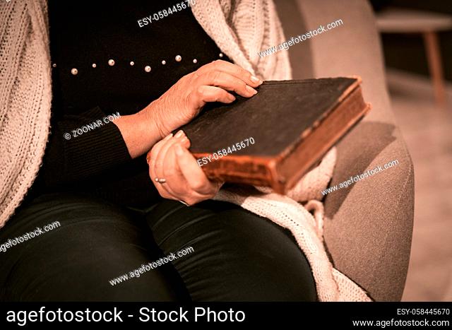 An Elderly Caucasian Woman Holds An Antique Book In Her Hands. Closed Book In An Old Dark Cover With Aged Pages. Selective Focus On Woman's Hands
