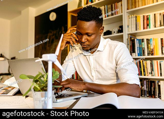 Male environmentalist talking on smart phone while using laptop at desk