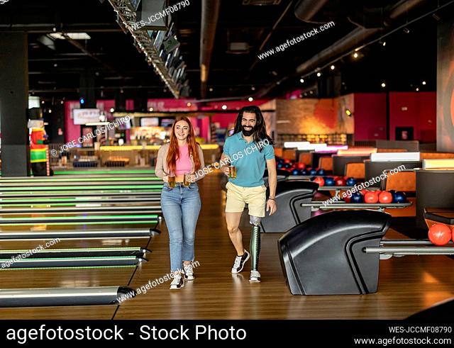 Happy man and woman with glasses of beer walking in bowling alley