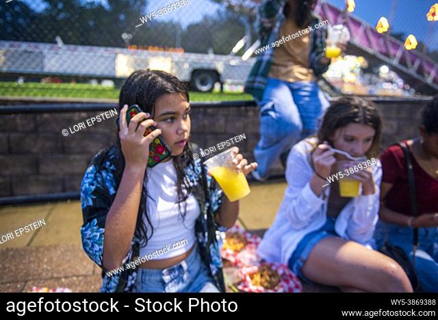A happy, diverse group of girl friends hang out at the county fair on a summer night. A hispanic girl talk on her cellphone while the others eat and drink and...