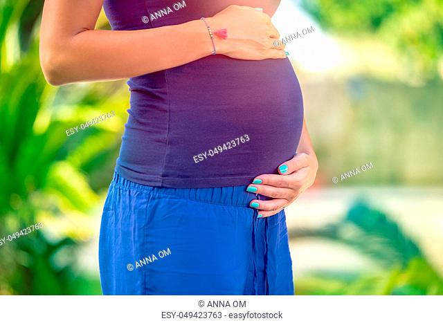 Closeup photo of a tummy of pregnant woman standing in the park, with gentleness and love future mother touching her belly, new life concept
