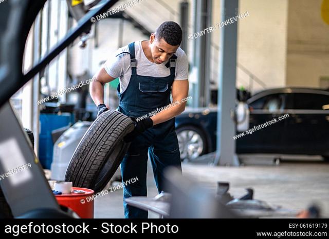 Young African American service station worker holding the rubber car wheel with the gloved hands