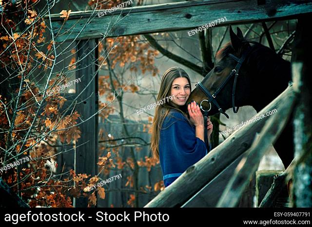 A beautiful girl in a blue stole stands next to a horse on the background of wooden buildings
