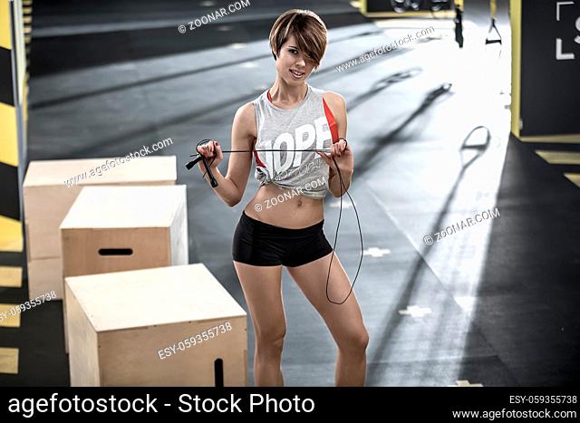 Athletic smiling girl with a skipping rope stands in the gym and looks into the camera. She wears a black shorts, red top and gray sleeveless