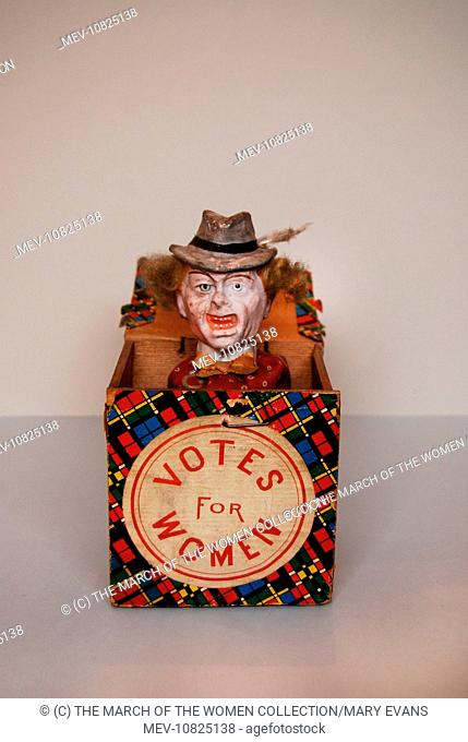 A suffragette Jack-in-the-Box with a bisque headed figure of a snarling woman. She has grey wiry hair which extends either side of a Tyrolean style hat with...