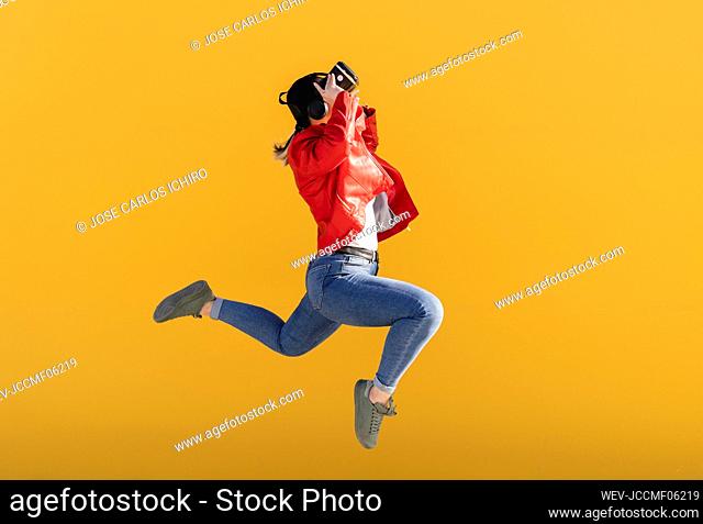 Playful woman wearing virtual reality simulator jumping against yellow background