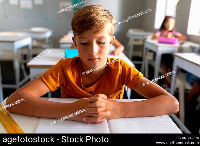 Caucasian elementary schoolboy with hands clasped studying at desk in classroom