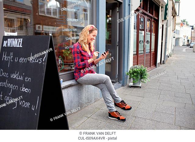 Woman sitting on shop windowsill looking down at smartphone smiling