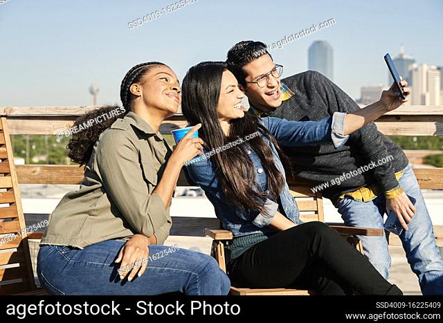 Group of young co-workers hanging out on rooftop patio, two women and man taking selfie with mobile phone