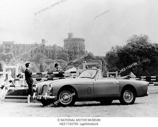 An Aston Martin DB2-4 MKII, with Windsor Castle in the background, 1956. An elegantly dressed woman and man stand behind the car in the middle of a show jumping...
