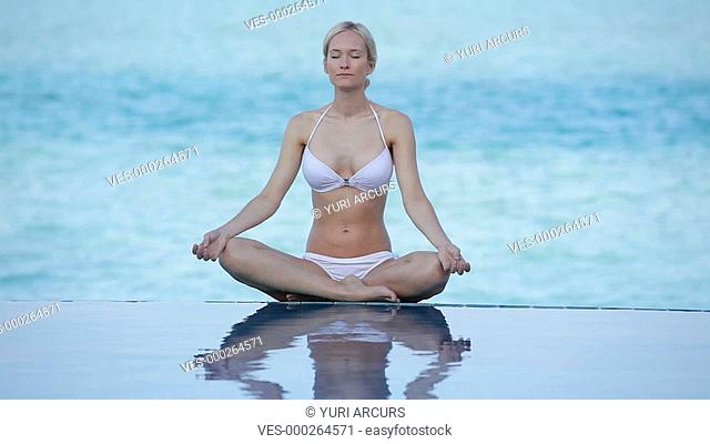 Pretty young woman sitting cross-legged meditating alongside a swimming-pool and the sea