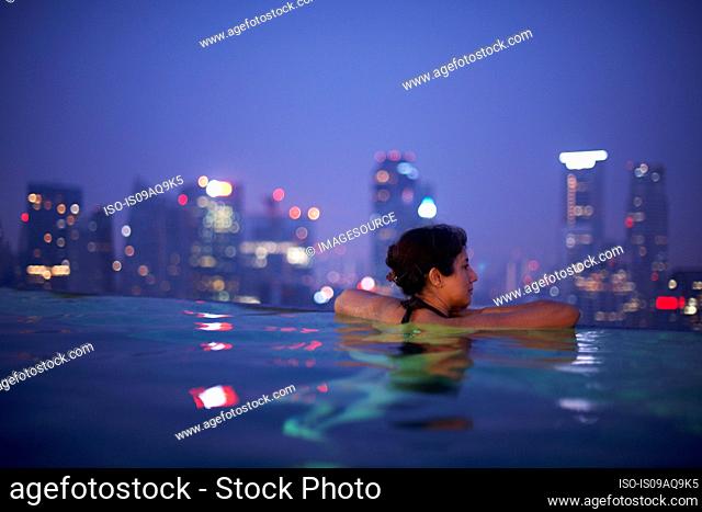 Mature female tourist looking out from hotel infinity pool at night, Singapore