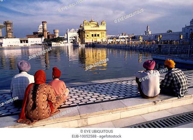 Sikh men, woman and child sitting on marble walkway beside the sacred pool looking towards the Golden Temple reflected in the water