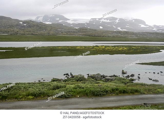 landscape with low clouds on green shore at lake in high valley of barren mountains with some summer snow, shot under bright cloudy summer light near Finse