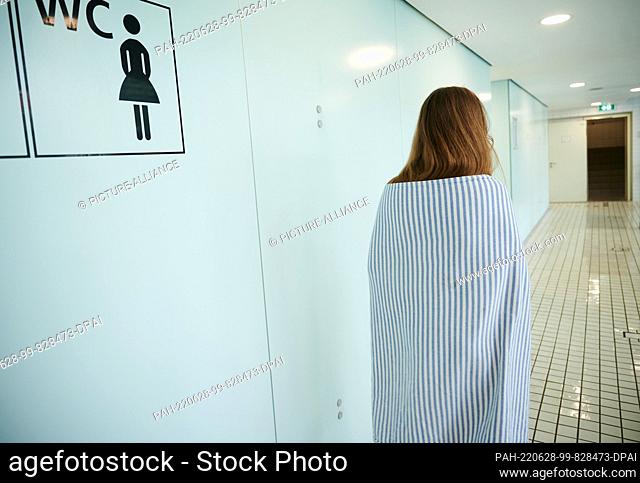 PRODUCTION - 25 June 2022, Berlin: ILLUSTRATION - A girl walks past a sign for a women's restroom in a swimming pool, covered with a towel