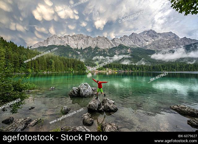 Young man jumps from rock to rock, Eibsee lake in front of Zugspitze massif with Zugspitze, dramatic Mammaten clouds, Wetterstein range, near Grainau