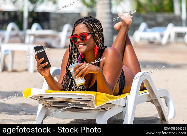 African-American woman on the beach relaxing in deck chair using laptop looking at camera. Cheerful adult woman enjoying a summer day at the beach