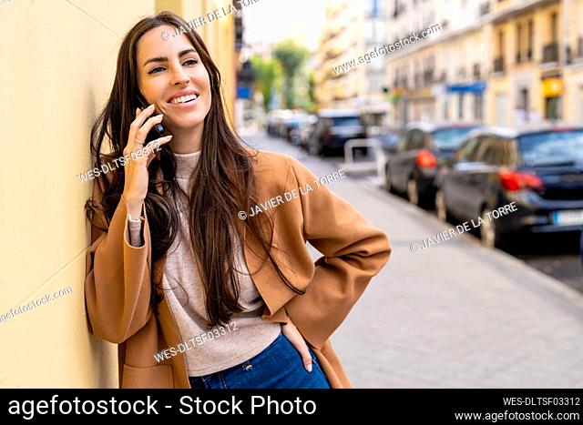 Smiling young woman talking over mobile phone leaning on yellow wall