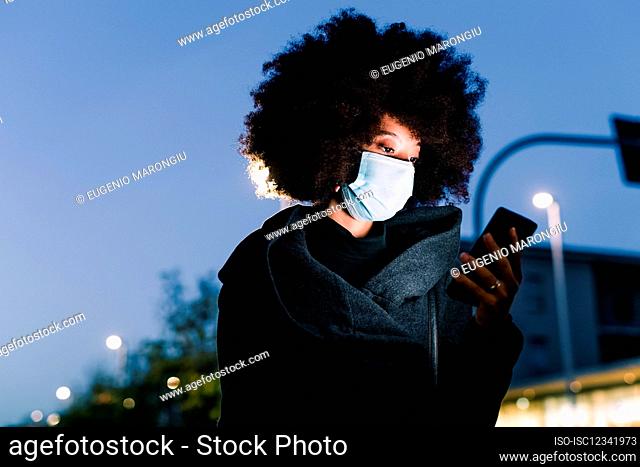 Woman wearing face mask and looking at phone, outdoors at night