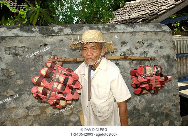 Half body portrait of an old man wearing white jacket sarong and a tattered straw hat standing and carrying bundles of red painted crude wooden sandals on a...