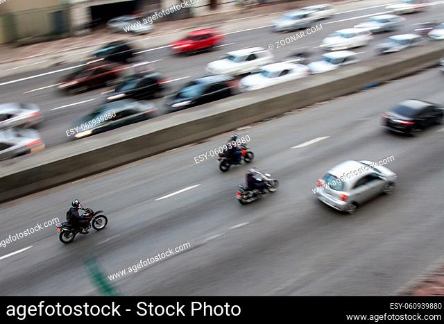 Pan shoot of motorcycles at high speed on an avenue in downtown from a nearby building with slower cars opposite direction lane. No people