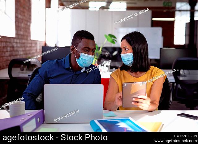 Diverse male and female colleague in face masks sitting at desk discussing, using laptop and tablet