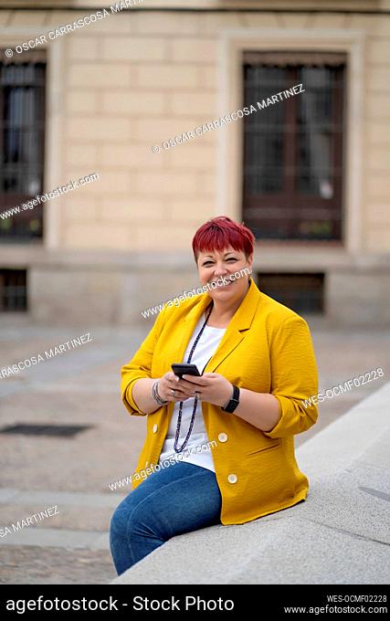 Smiling woman with mobile phone sitting on retaining wall