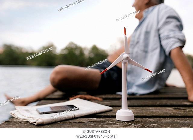 Senior man sitting on jetty at a lake with small wind turbine model